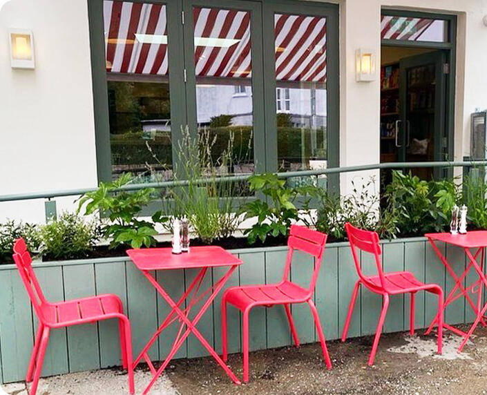 Pink metal chairs and small tables on the terrace, behind a planter and under a striped awning