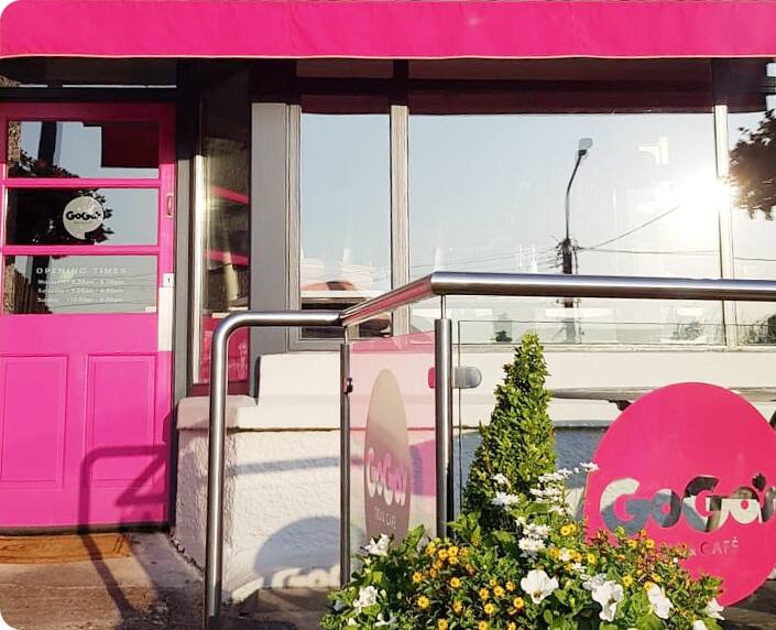 The pink door, awning and round GoGo's sign at the front of the shop, with a planter of white and yellow flowers